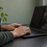 Two hands at a dark unbranded laptop with a blank reflective screen in a daylight workspace